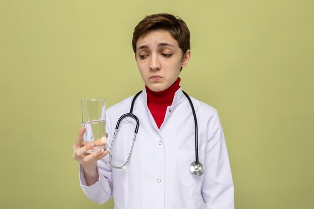 Jonge vrouwelijke arts in witte jas met stethoscoop om nek die glas water vasthoudt en er verward naar kijkt terwijl hij op groen staat
