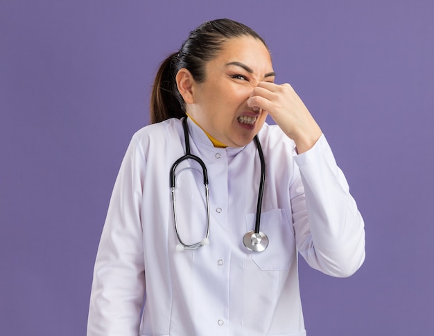 Jonge vrouwelijke arts in witte medicijnjas met stethoscoop om nek die neus sluit met vingers die lijden aan stank die over paarse muur staat