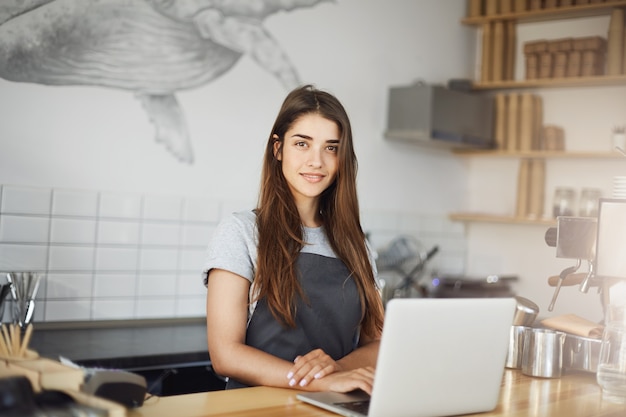 Jonge vrouwelijke barista met behulp van een laptopcomputer op haar werk in café. Gelukkige werknemer die camera het glimlachen bekijkt.