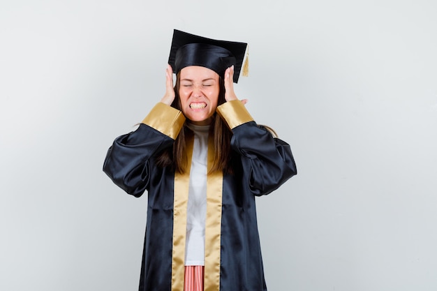 Jonge vrouwelijke gediplomeerde die handen op agressieve manier in geïsoleerde academische kleding houdt