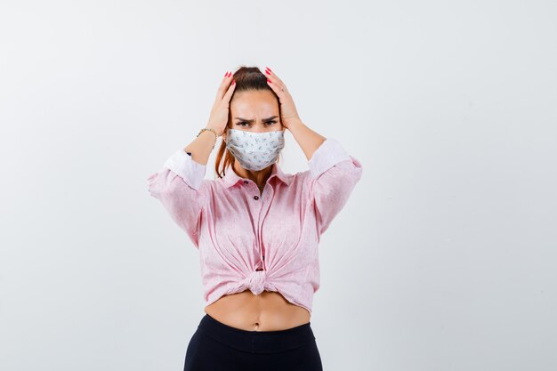 Jonge vrouwelijke hand in hand op het hoofd in shirt, broek, medisch masker en op zoek naar vergeetachtig, vooraanzicht.