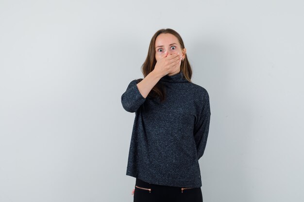 Jonge vrouwelijke hand op de mond in zwarte blouse en bang op zoek. vooraanzicht.