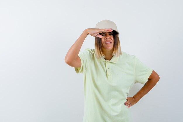 Jonge vrouwelijke hand op het hoofd vast te houden om duidelijk te zien terwijl de hand op de taille in t-shirt, pet en gefocust, vooraanzicht.