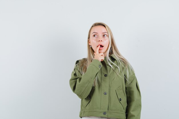 Jonge vrouwelijke holdingsvinger op haar lippen die in groen jasje denken en peinzend, vooraanzicht kijken.