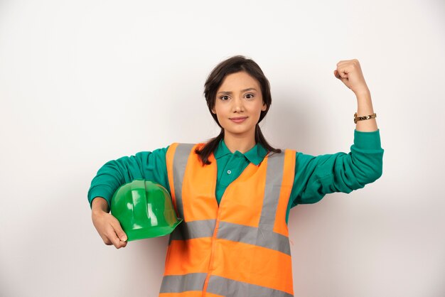 Jonge vrouwelijke ingenieur die spieren toont en een helm op witte achtergrond houdt