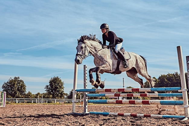 Jonge vrouwelijke jockey op vlek grijs paard springen over hindernis in de open arena.