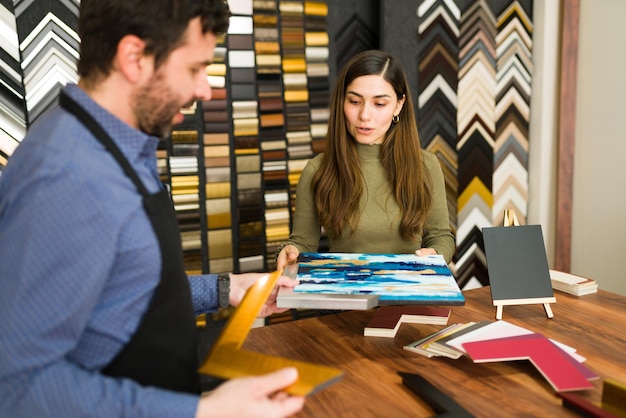 Jonge vrouwelijke klant in de winkel probeert verschillende frames voor haar kleine canvas en praat met een aantrekkelijke mannelijke werknemer