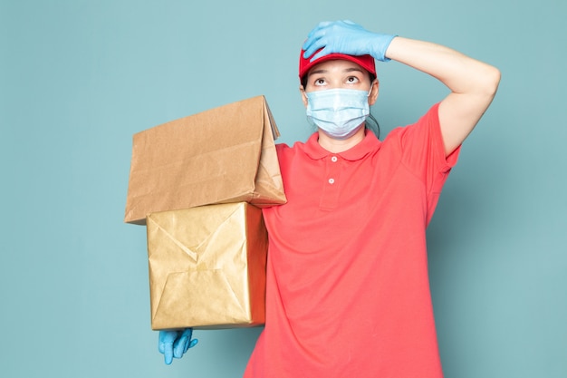 jonge vrouwelijke koerier in roze t-shirt rode dop blauw steriel masker blauwe handschoenen met doos op de blauwe muur