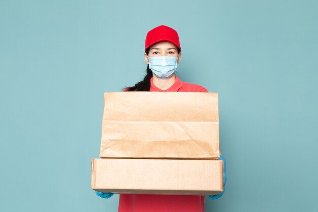 jonge vrouwelijke koerier in roze t-shirt rode dop blauw steriel masker blauwe handschoenen met doos op de blauwe muur