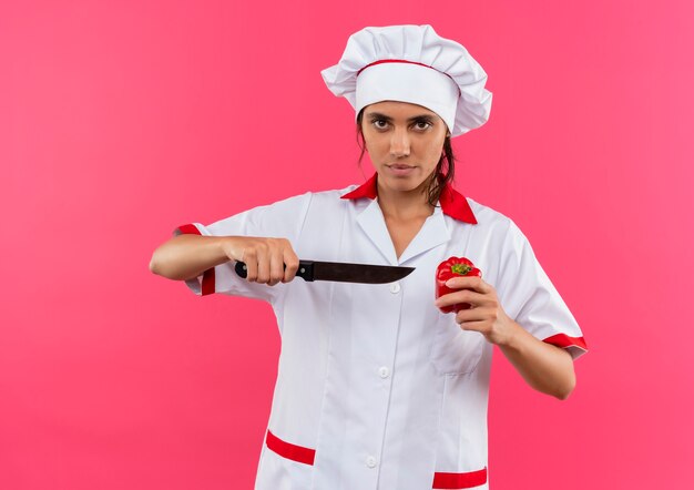 jonge vrouwelijke kok die chef-kok het uniforme mes en peper op geïsoleerde roze muur met exemplaarruimte draagt