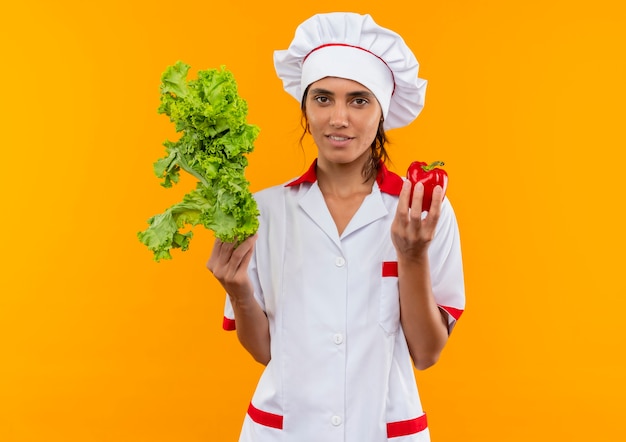jonge vrouwelijke kok die de salade en de peper van de chef-kok uniforme op geïsoleerde gele muur met exemplaarruimte draagt