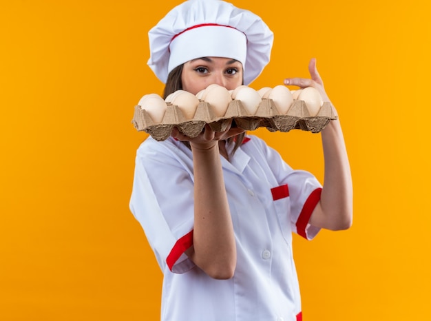 Jonge vrouwelijke kok die een uniform van de chef-kok draagt en wijst naar een partij eieren geïsoleerd op een oranje muur