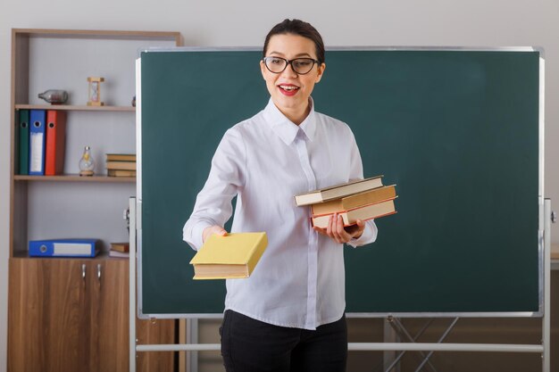 Jonge vrouwelijke leraar die een bril draagt en uitleg geeft over de les die een stapel boeken vasthoudt die glimlacht, zelfverzekerd staat aan de schoolbank voor het bord in de klas