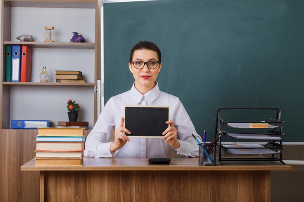 Jonge vrouwelijke leraar die een bril draagt met een klein schoolbord glimlachend zelfverzekerd zittend aan de schoolbank voor het bord in de klas