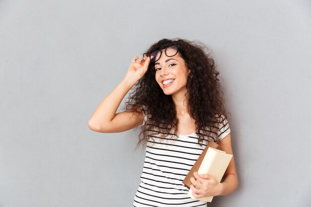 Jonge vrouwelijke leraar in oogglazen met krullend haar die zich met boeken in hand over grijze muur bevinden die van haar werk in universiteit genieten die slim en intellectueel zijn