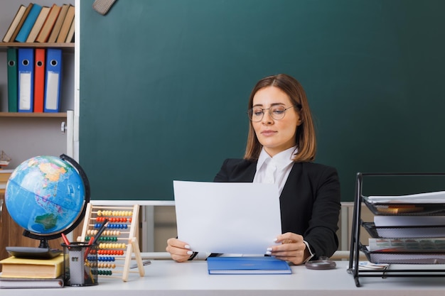 Jonge vrouwelijke leraar met een bril die aan de schoolbank zit met globe en boeken voor het bord in de klas met een wit leeg vel papier dat er zelfverzekerd uitziet