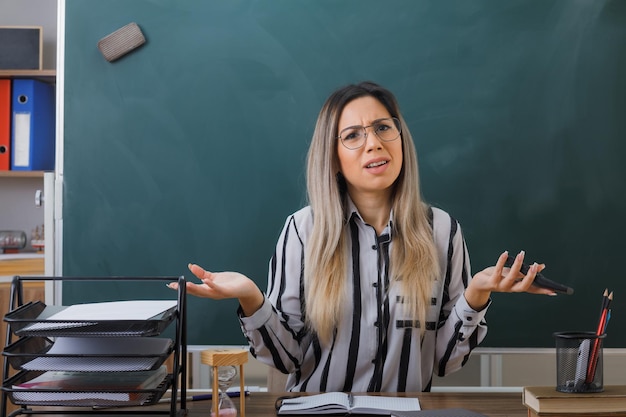 jonge vrouwelijke leraar met een bril die aan de schoolbank zit voor het bord in de klas en uitleg geeft over de les die er verward en teleurgesteld uitziet