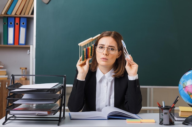 Jonge vrouwelijke leraar met een bril die aan de schoolbank zit voor het bord in de klas met nummerplaten en telraam die de les uitleggen en er verbaasd uitzien