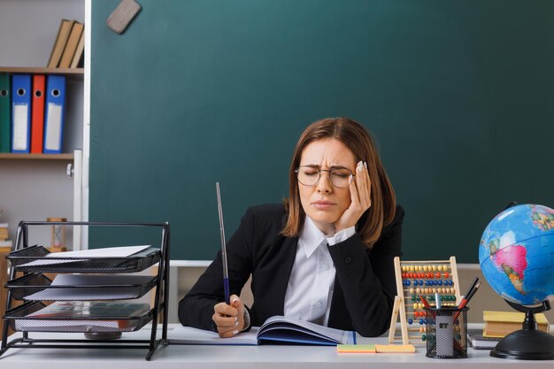 Jonge vrouwelijke leraar met een bril die aan de schoolbank zit voor het schoolbord in de klas met telraam en wereldbol die klassenregister controleert met aanwijzer die er onwel uitziet, pijn voelt