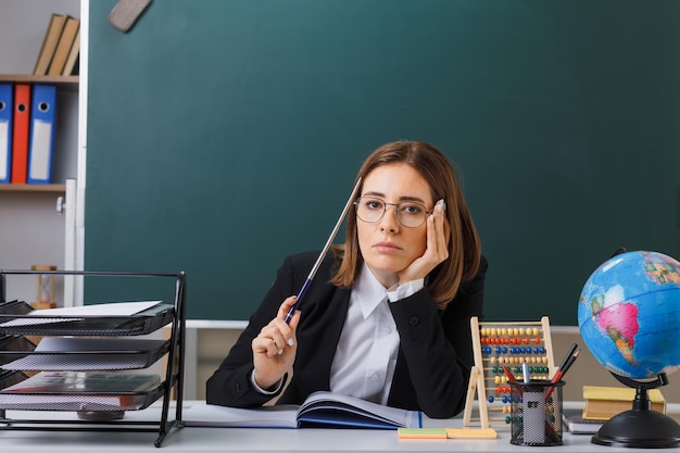 Jonge vrouwelijke leraar met een bril die aan een schoolbank zit voor een schoolbord in de klas met een telraam en een wereldbol die het klassenregister controleert met de aanwijzer die er moe en verveeld uitziet