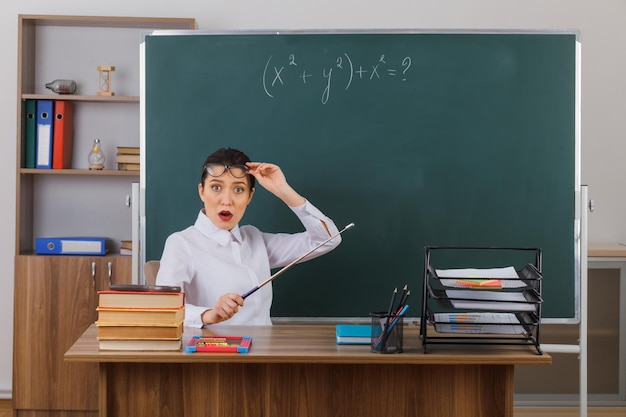 Jonge vrouwelijke leraar met een bril die de aanwijzer vasthoudt terwijl hij de les uitlegt die er verrast en geschokt uitziet terwijl hij aan de schoolbank voor het bord in de klas zit
