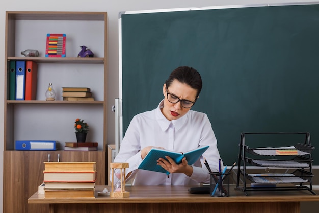 Jonge vrouwelijke leraar met een bril die een boek leest met een serieus gezicht dat zich voorbereidt op de les die aan de schoolbank zit voor het bord in de klas