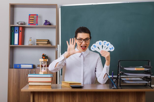 Jonge vrouwelijke leraar met een bril die nummerplaten vasthoudt en les uitlegt met nummer vijf met open palm gelukkig en vrolijk zittend aan de schoolbank voor het bord in de klas