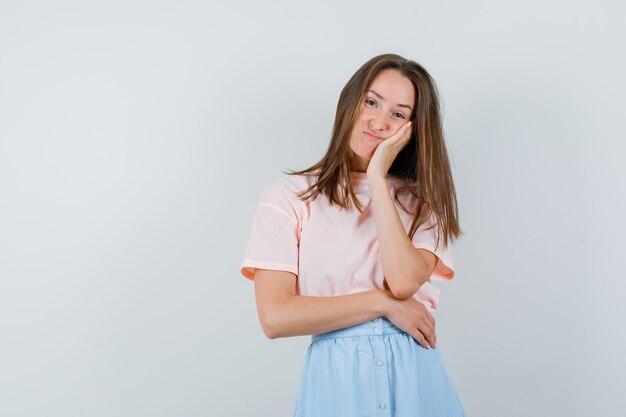 Jonge vrouwelijke leunende wang op opgeheven palm in t-shirt, rok en op zoek teleurgesteld, vooraanzicht.