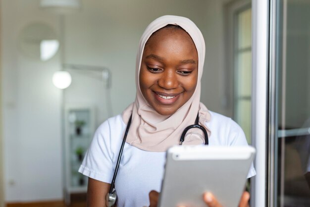 Jonge vrouwelijke moslim Afrikaanse clinicus arts in scrubs met behulp van touchpad tijdens online communicatie met patiënten