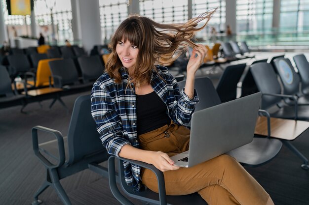 Jonge vrouwelijke passagier met laptop zittend in terminal hal tijdens het wachten op haar vlucht