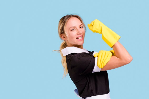 Jonge vrouwelijke reinigingsmachine die haar spier toont tegen blauwe muur