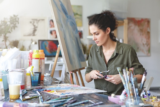 Jonge vrouwelijke schilder zittend aan tafel omringd met verschillende penselen en aquarellen terwijl het creëren van mooie foto op ezel. Creatieve werknemer bezig met canvas. Ambacht en kunstconcept