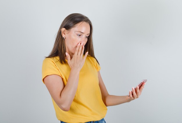 Jonge vrouwelijke smartphone met hand op mond in geel t-shirt, korte broek en op zoek naar verrassing te houden.
