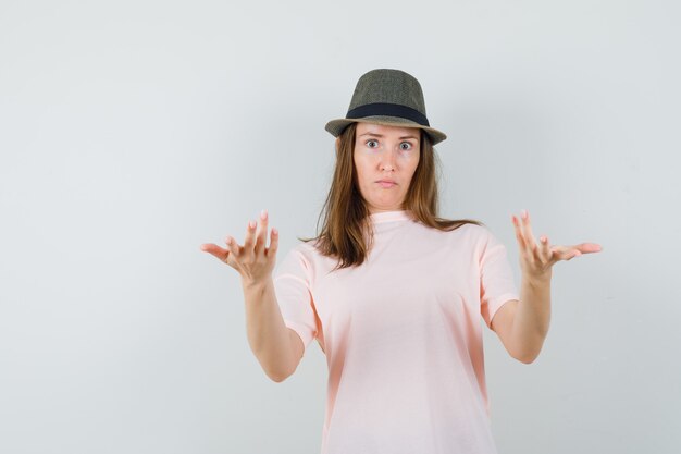 Jonge vrouwelijke strekken handen in verbaasd gebaar in roze t-shirt, hoed vooraanzicht.