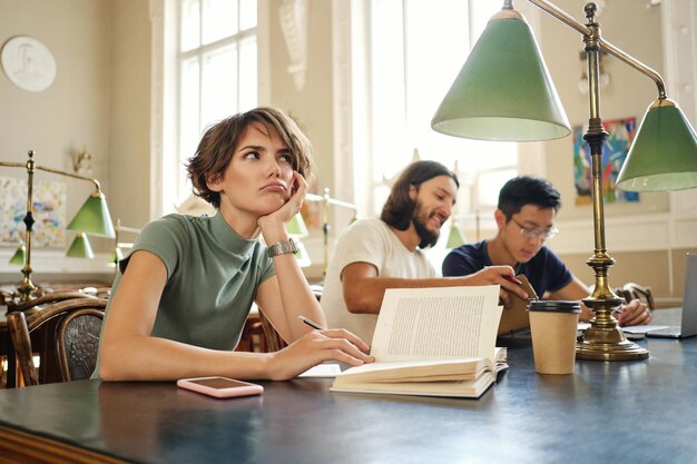 Jonge vrouwelijke student met boek die zorgvuldig aan nieuw studieproject in bibliotheek van universiteit werkt