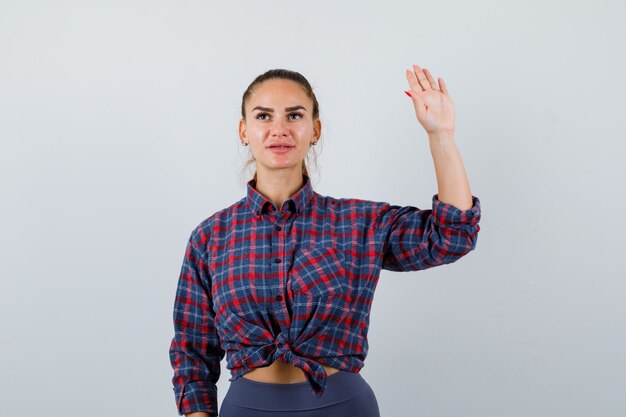 Jonge vrouwelijke zwaaiende hand voor begroeting in geruit hemd, broek en zelfverzekerd, vooraanzicht.