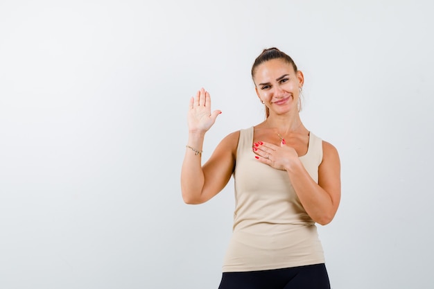 Jonge vrouwelijke zwaaiende hand voor groet in beige mouwloos onderhemd en op zoek gelukkig, vooraanzicht.