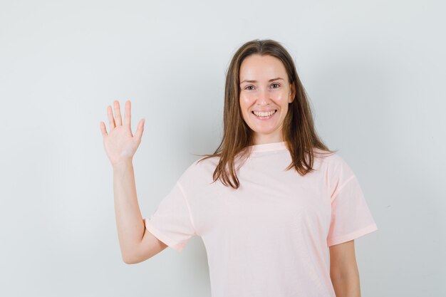 Jonge vrouwelijke zwaaiende hand voor groet in roze t-shirt en op zoek vrolijk. vooraanzicht.