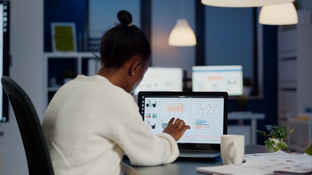 Jonge vrouwenmanager die bedrijfsstatistieken en financiële rapporten controleert op laptop die 's avonds laat in een opstartkantoor werkt