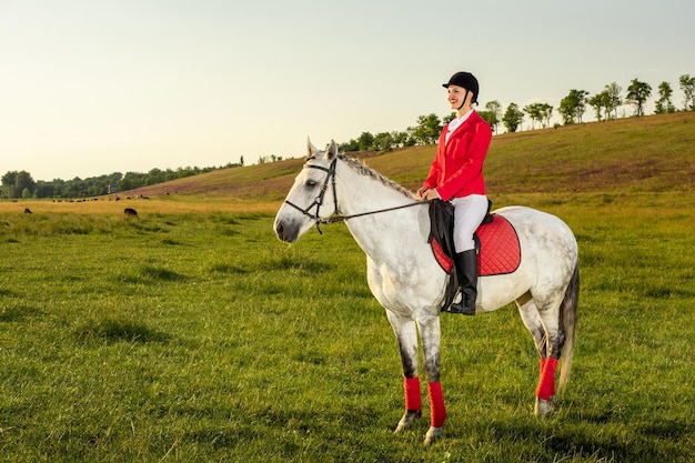 Jonge vrouwenruiter, die rode redingote en witte rijbroek draagt, met haar paard in avondzonsonderganglicht.