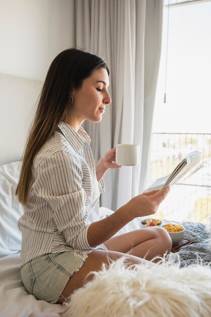 Jonge vrouwenzitting op de krant van de bedlezing terwijl het drinken van koffie