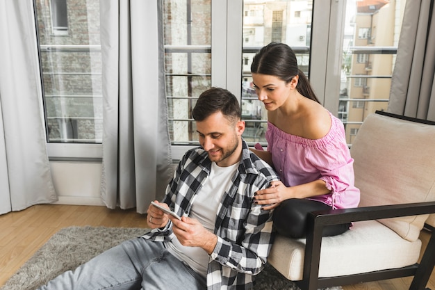 Jonge vrouwenzitting op stoel die haar vriend thuis bekijkt die mobiele telefoon met behulp van