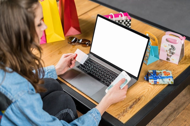 Jonge vrouwenzitting voor laptop met het lege scherm die mobiele telefoon en creditcard in hand houden