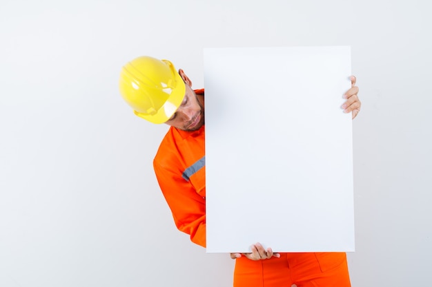 Jonge werknemer die leeg canvas in uniform, helm bekijkt en gericht kijkt.