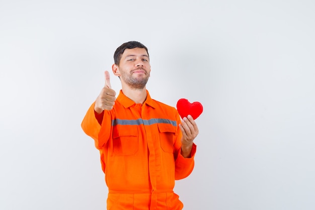 Jonge werknemer die rood hart houdt, duim in uniform toont en tevreden kijkt.