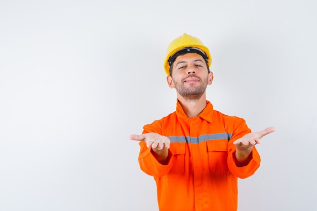 Jonge werknemer handen strekken als iets aanbieden in uniform, helm en er vrolijk uitzien.