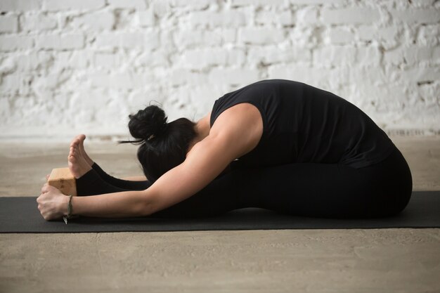 Jonge yogi vrouw in paschimottanasana poseren met blok, loft terug