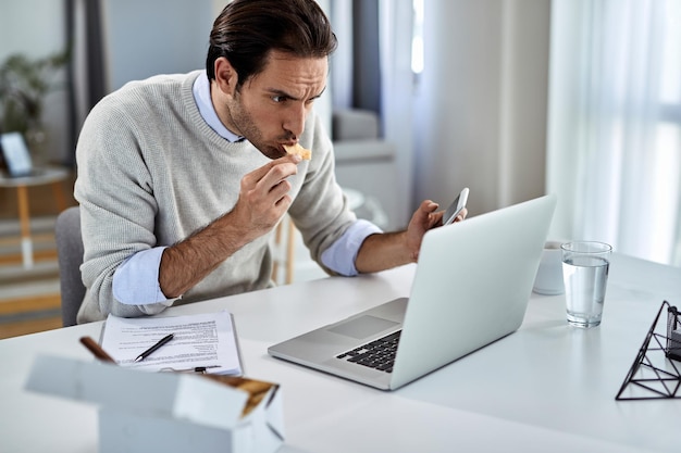 Jonge zakenman die eet terwijl hij een mobiele telefoon gebruikt en thuis op een laptop werkt