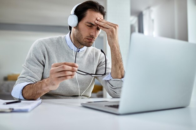 Jonge zakenman die hoofdpijn heeft en zich uitgeput voelt terwijl hij thuis op een laptop werkt