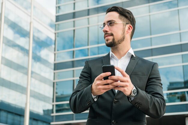 Jonge zakenman die zich voor de bureaubouw bevindt die mobiele telefoon houdt